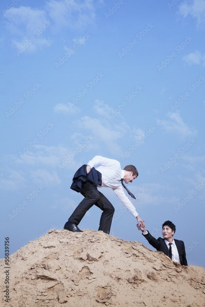 Young businessman helping another businessman get to the top of the hill in the desert Young businessman helping another businessman get to the top of the hill in the desert