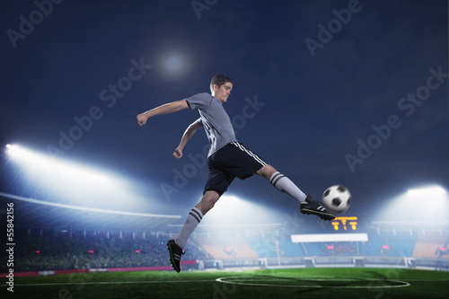 Soccer player in mid air kicking the soccer ball, stadium lights at night in background © xixinxing