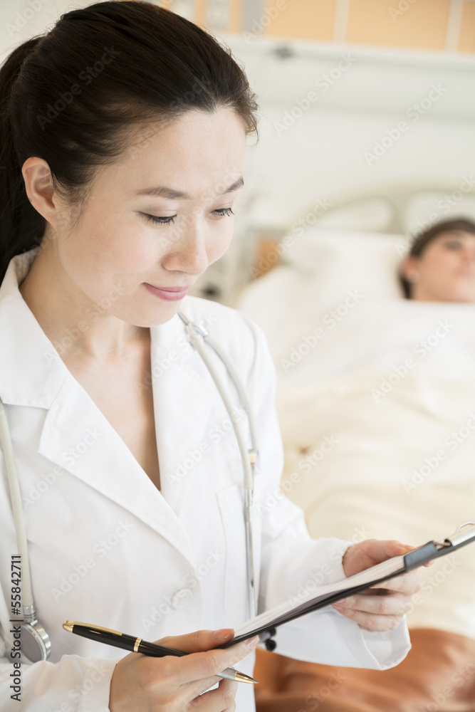 Portrait of smiling doctor holding a medical chart with patient lying in a hospital bed in the background Portrait of smiling doctor holding a medical chart with patient lying in a hospital bed in the background