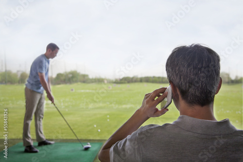 Rear view of man on the phone while another man plays golf in the background