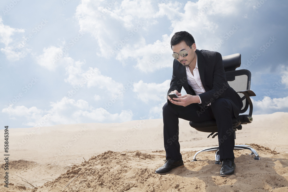 Young businessman in sunglasses sitting in an office chair in the middle of the desert, checking his phone Young businessman in sunglasses sitting in an office chair in the middle of the desert, checking his phone