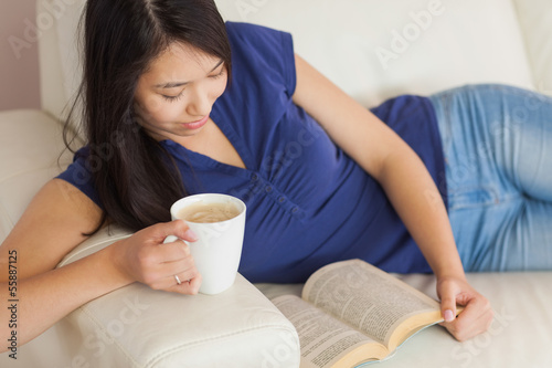 Happy young asian woman lying on the sofa reading a book holding