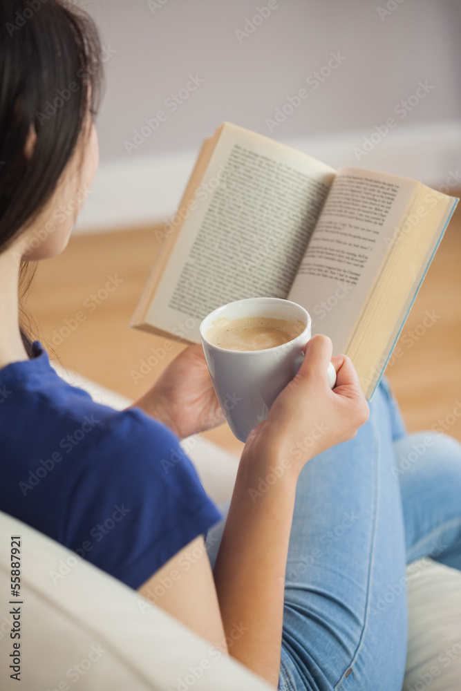 Obraz premium Young woman sitting on the sofa reading a book holding her coffe
