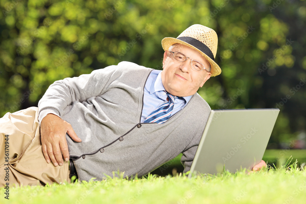 Fototapeta premium Senior man with hat lying on a grass working on a laptop in park