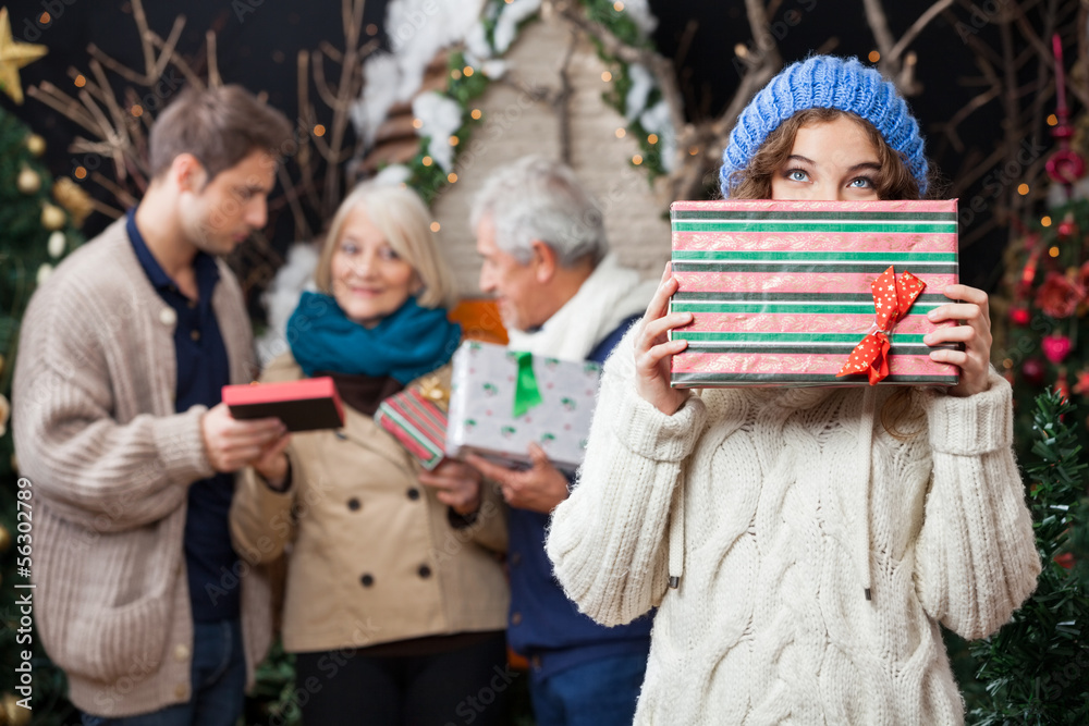 Thoughtful Woman Holding Christmas Present With Family In Backgr