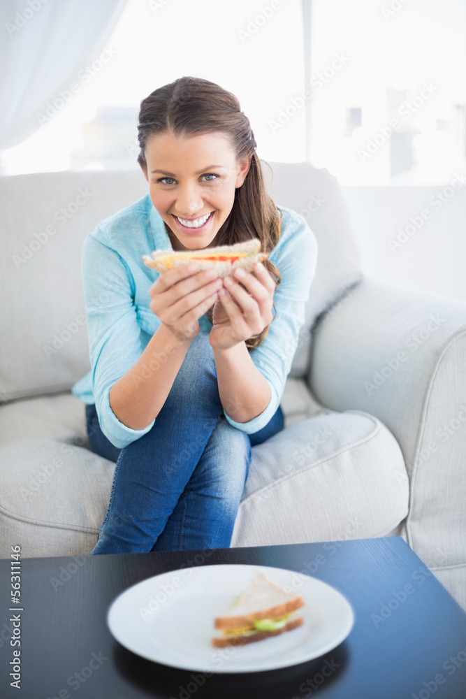 Happy woman sitting on sofa showing sandwich at camera Happy woman sitting on sofa showing sandwich at camera