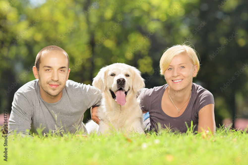 Smiling young couple lying on a grass and hugging a dog in a par Smiling young couple lying on a grass and hugging a dog in a par