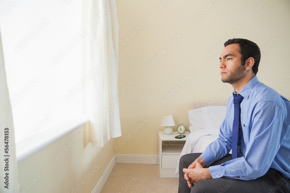 Unsmiling man looking out the window sitting on his bed