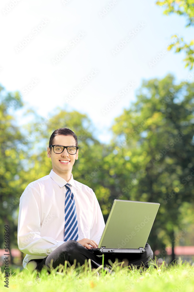 Smiling businessman seated on a grass with laptop in a park Smiling businessman seated on a grass with laptop in a park