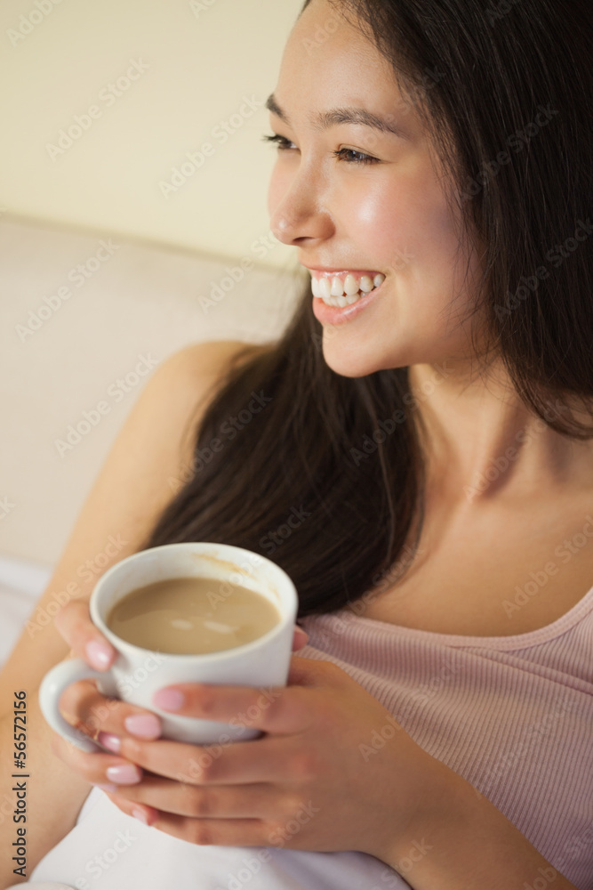 Cheerful young asian woman sitting in bed holding her morning co Cheerful young asian woman sitting in bed holding her morning co