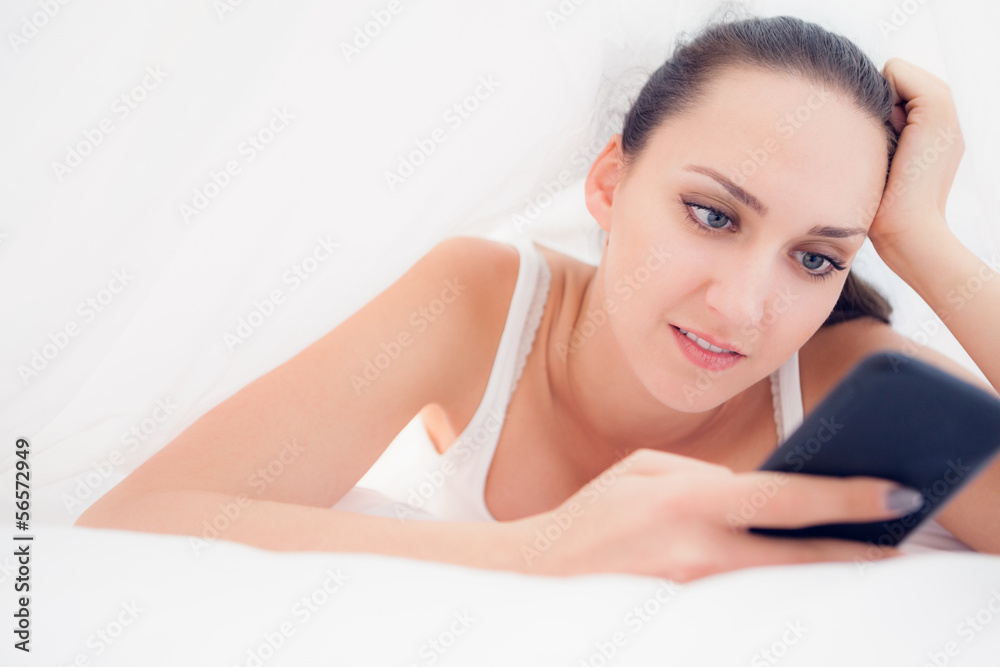Happy brunette lying under the sheets using her smartphone Happy brunette lying under the sheets using her smartphone