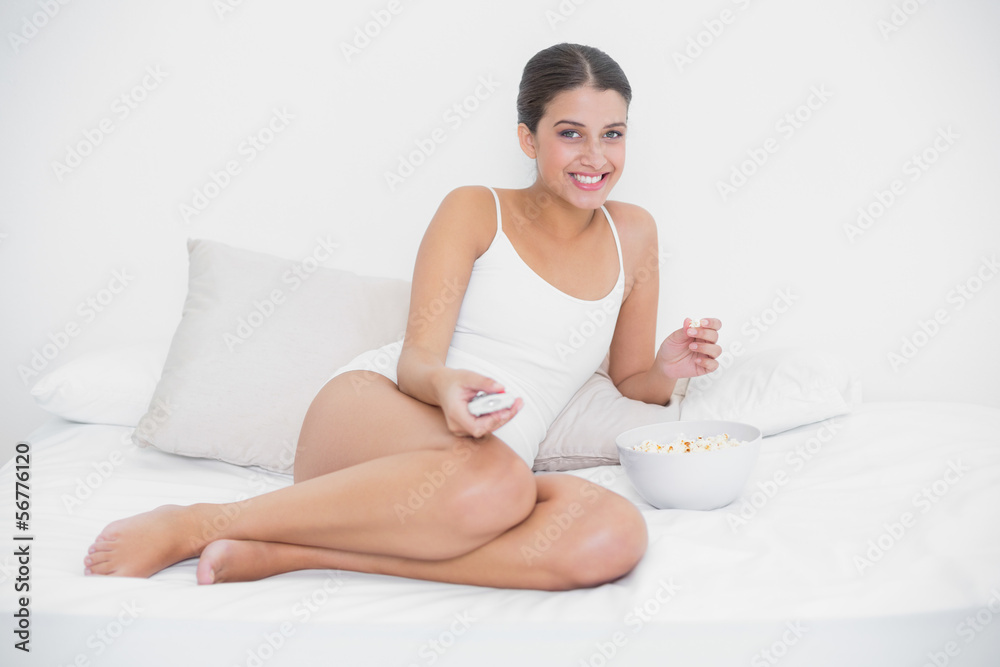 Amused young brown haired model in white pajamas watching tv whi Amused young brown haired model in white pajamas watching tv whi