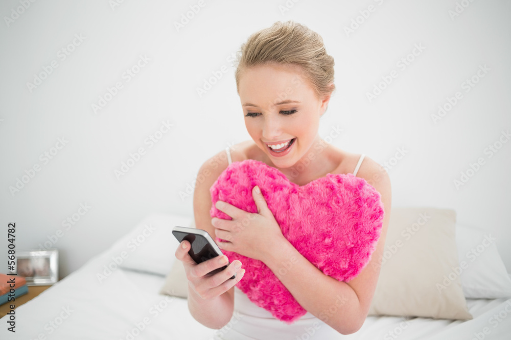 Natural smiling blonde holding heart pillow and smartphone Natural smiling blonde holding heart pillow and smartphone