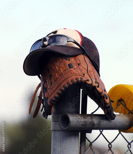 Baseball cap, mitt and sunglasses on fence at field.