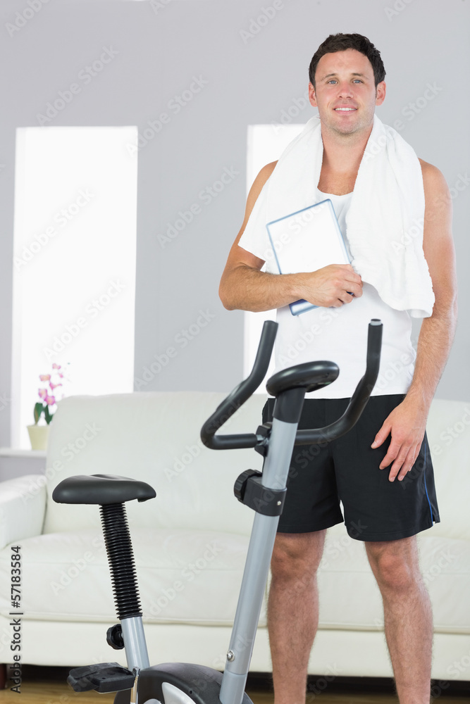 Cheerful sporty man standing behind exercise bike holding tablet Cheerful sporty man standing behind exercise bike holding tablet