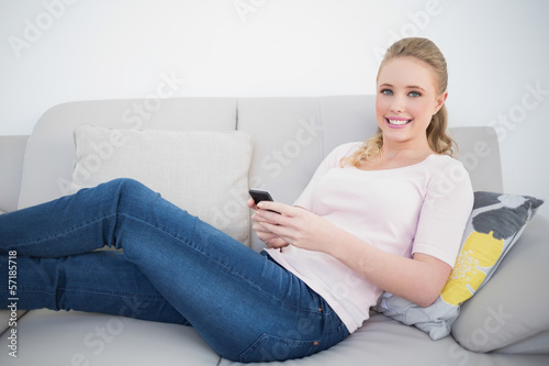 Casual cheerful blonde using smartphone and lying on couch