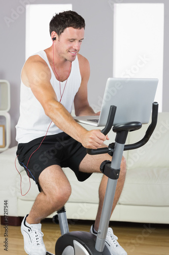 Sporty man with earphones exercising on bike looking at laptop