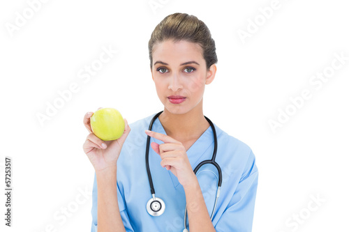 Stern brown haired nurse in blue scrubs pointing an apple with h