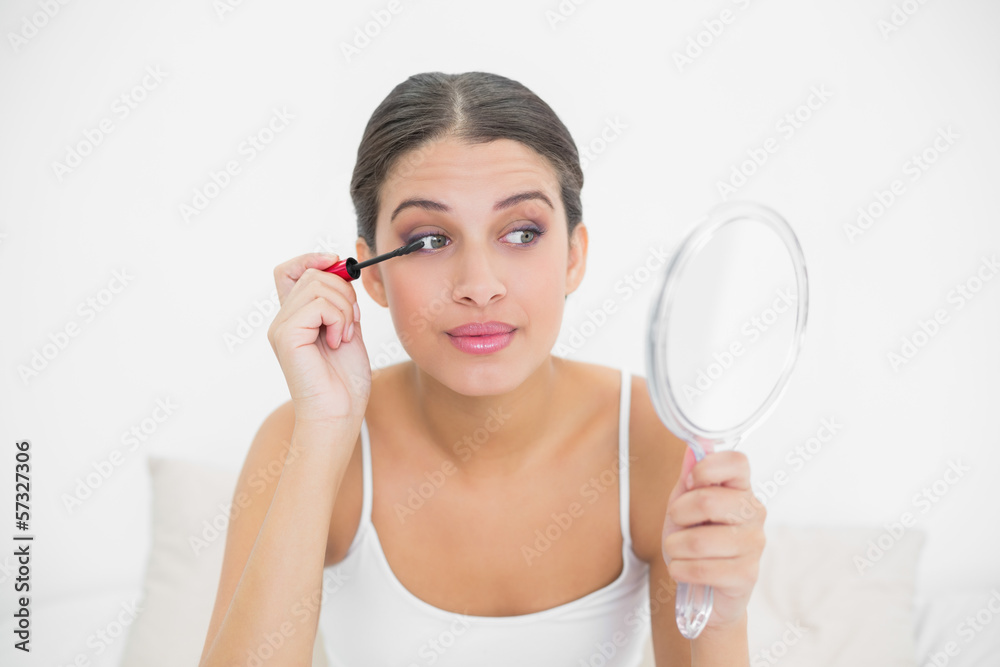 Focused young brown haired model in white pajamas applying masca Focused young brown haired model in white pajamas applying masca