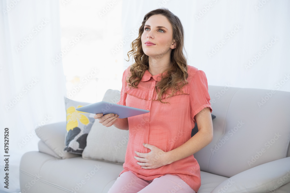 Thinking pregnant woman holding her tablet while sitting on a co Thinking pregnant woman holding her tablet while sitting on a co