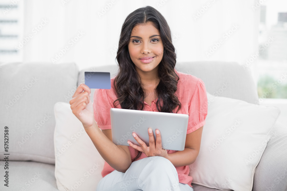 Pleased cute brunette sitting on couch holding credit card and t Pleased cute brunette sitting on couch holding credit card and t