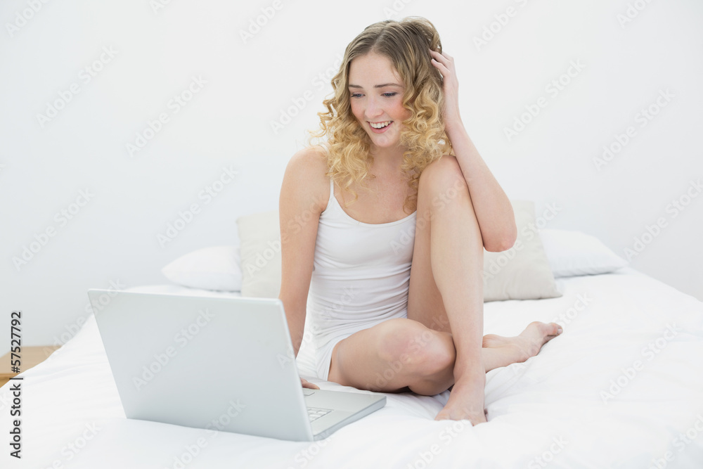 Pretty smiling blonde sitting on bed using laptop Pretty smiling blonde sitting on bed using laptop