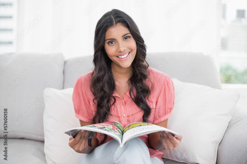 Smiling cute brunette sitting on couch holding magazine Smiling cute brunette sitting on couch holding magazine