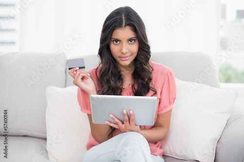 Calm cute brunette sitting on couch holding credit card and tabl