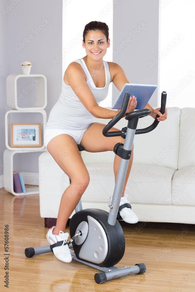 Happy training woman using an exercise bike while holding a tabl Happy training woman using an exercise bike while holding a tabl