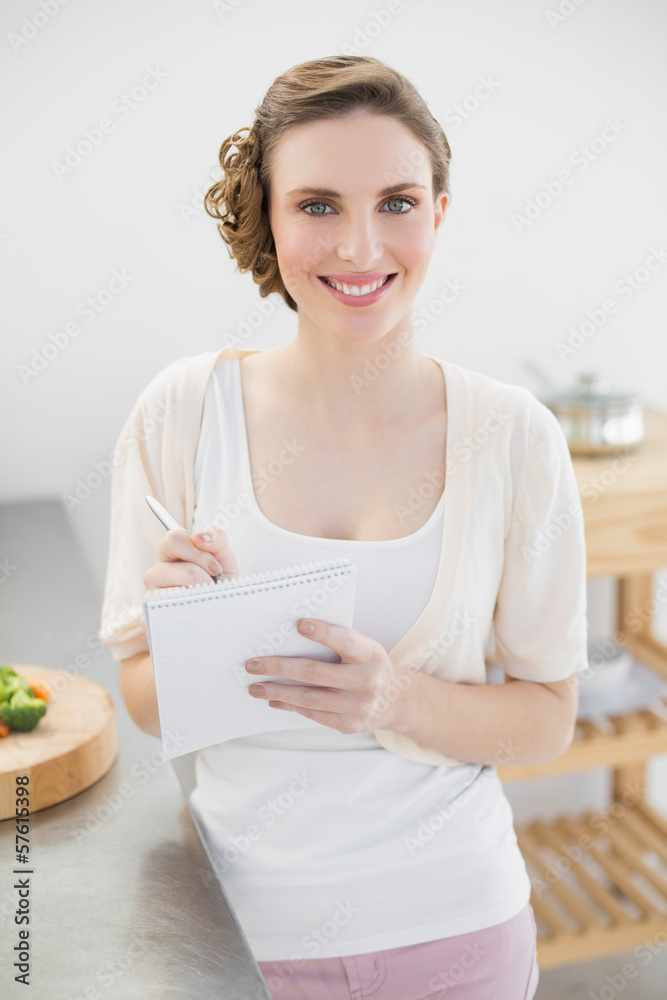 Young woman standing in her kitchen writing a shopping list smil