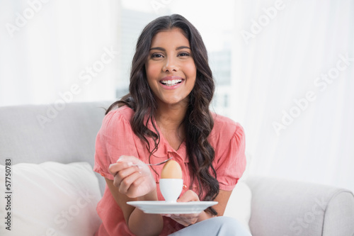 Cheerful cute brunette sitting on couch holding hard boiled egg