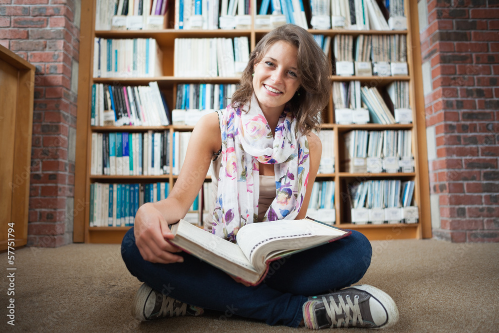 Happy female student against bookshelf with a book on the librar Happy female student against bookshelf with a book on the librar