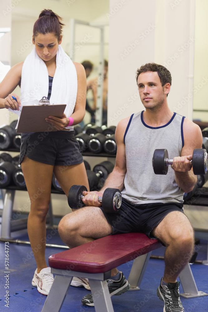 Trainer taking notes of muscular man lifting dumbbells Trainer taking notes of muscular man lifting dumbbells