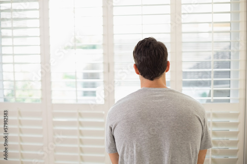 Rear view of man looking through window blinds at bright room