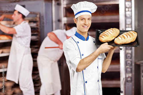 Leinwand Poster Smiling male baker posing with freshly baked breads in bakery