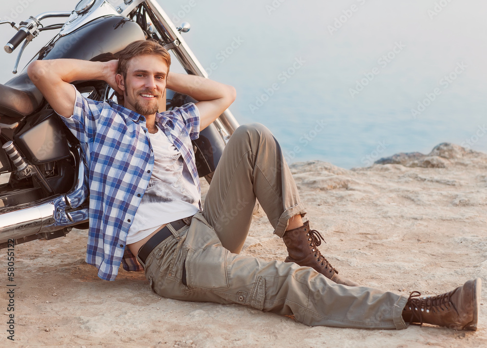 Man is sitting on the beach with his back to the bike.