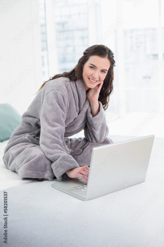 Smiling brunette in bathrobe using laptop in bed Smiling brunette in bathrobe using laptop in bed