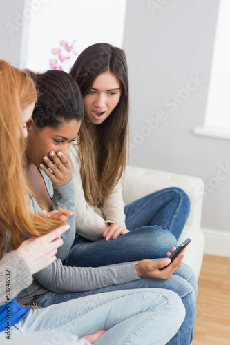 Shocked friends looking at mobile phone together on sofa