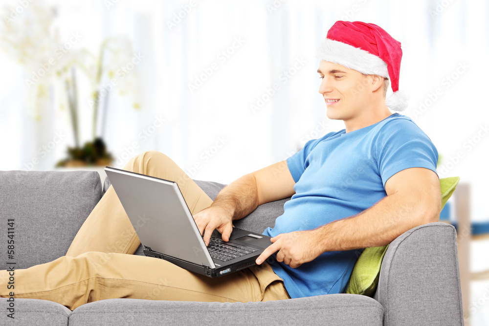 Handsome guy with santa hat on a sofa working on a laptop home Handsome guy with santa hat on a sofa working on a laptop home