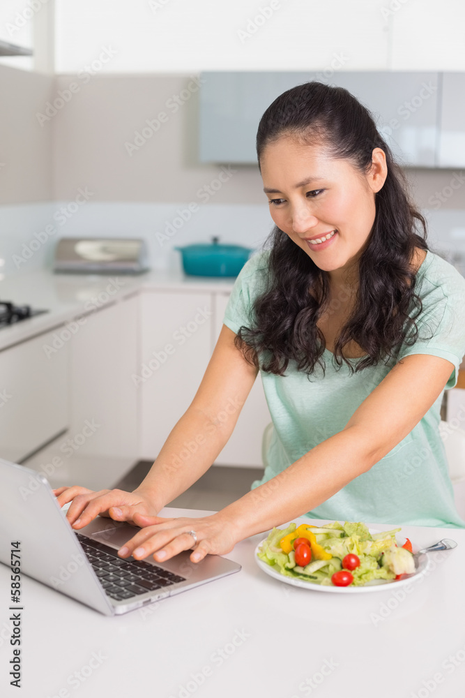 Smiling woman using laptop while having salad in kitchen