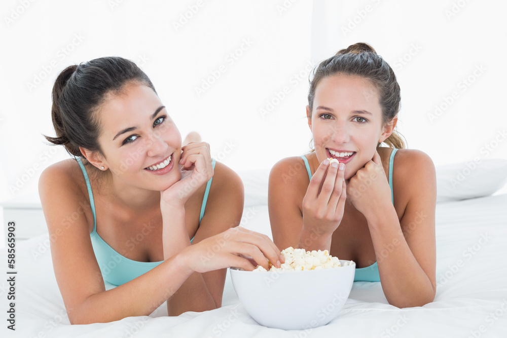 Smiling female friends eating popcorn in bed Smiling female friends eating popcorn in bed