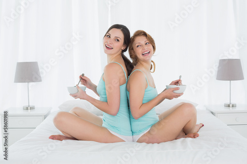Cheerful female friends with salad bowls sitting on bed