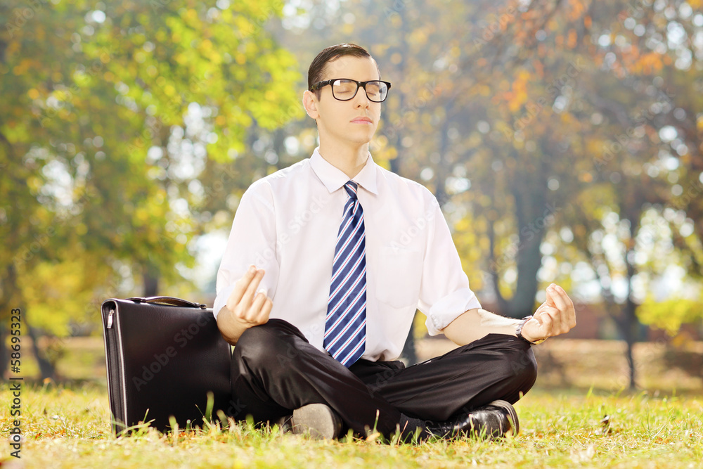 Young businessman seated on a green grass meditating in a park Young businessman seated on a green grass meditating in a park