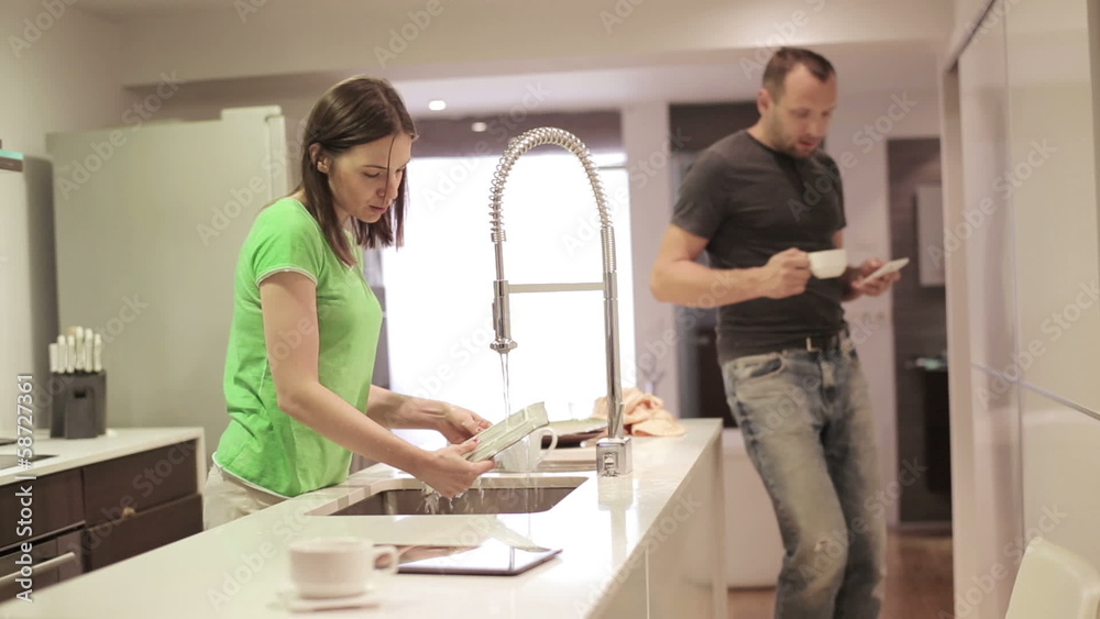 Angry woman washing dishes while man texting on smartphone Angry woman washing dishes while man texting on smartphone