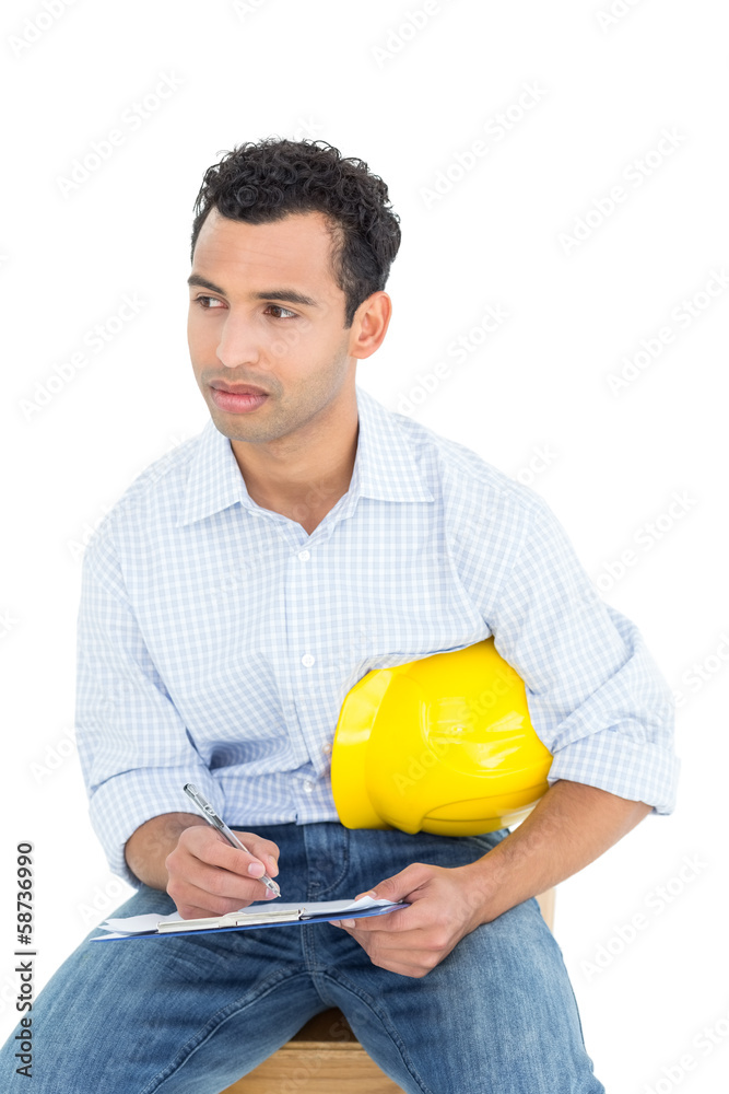 Thoughtful handyman with yellow hard hat writing in clipboard
