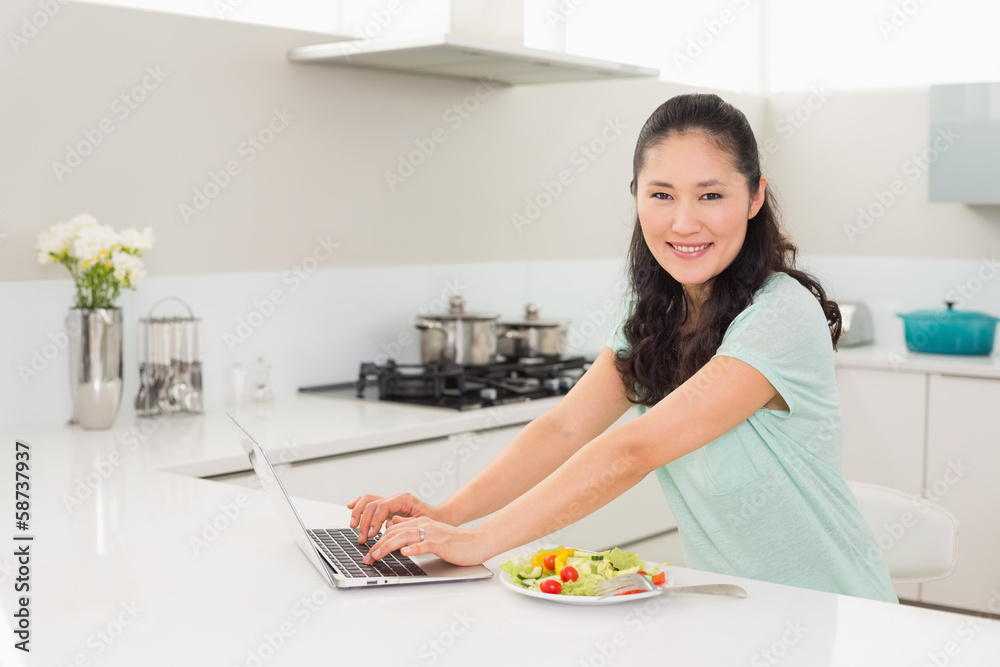 Woman using laptop while having salad in kitchen