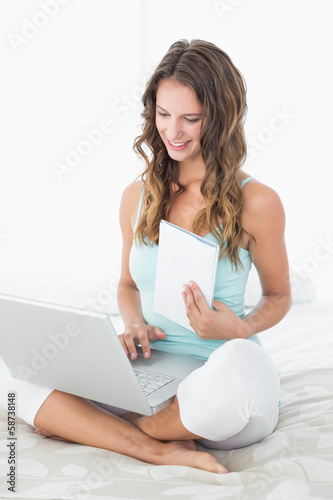Woman using laptop as she holds a notepad in bed
