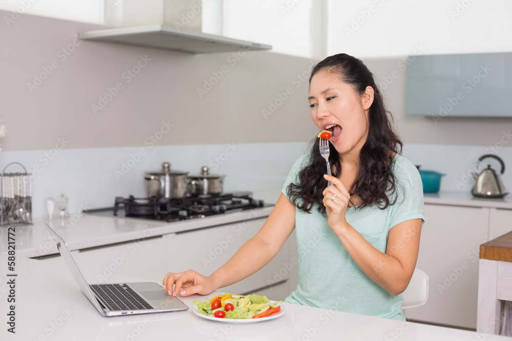 Young woman with laptop eating salad in kitchen Young woman with laptop eating salad in kitchen