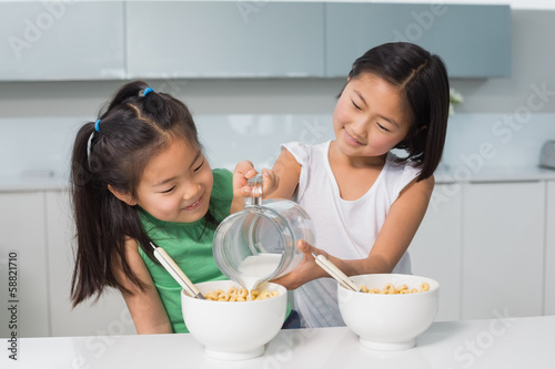 Two happy young girls pouring milk in bowl in kitchen