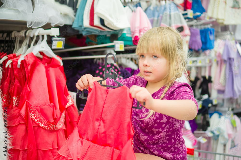 Adorable girl on shoppping cart select red dress in supermarket